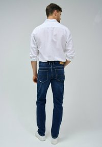 Vaqueros de mezclilla azul oscuro con un corte ajustado, detalles de costura visibles y dos bolsillos traseros. Se combinan con una camisa blanca y zapatillas blancas.
