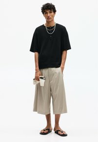Chemise noire à manches courtes texturée avec des boutons, pantalon large beige clair, et tongs noires ; tenant un petit sac tissé blanc.