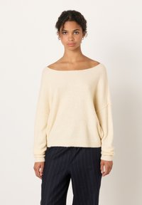 Sieviete valkā krēmkrāsas džemperi ar atklātiem pleciem un tumšas ar svītrām bikses, stāvot pret vienkāršu gaišu fonu.