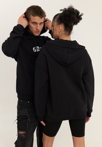 Dva modely nosia pre veľké čierne mikiny s predným vreckom, ktoré majú plochú textúru a jemný detail loga. Vidno aj čierne roztrhnuté džínsy.