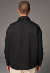 Sort langærmet skjorte med krave, der viser en struktureret stof med en afslappet pasform og sideslids ved kanten. Synlige syninge detaljer.