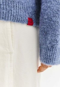 Jasnoniebieski dziergany sweter o prążkowanej fakturze z czerwoną metką, noszony na białych spodniach z widocznym detalem kieszeni.