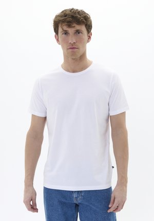 T-shirt bianco a maniche corte con scollo rotondo, realizzato in cotone morbido, abbinato a jeans in denim blu, con un'etichetta del logo sul lato.