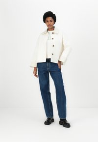 Cream-colored cropped jacket with large buttons, worn over a light sweatshirt with green text, paired with blue straight-leg jeans and black shoes.