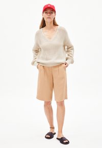 Cream knitted V-neck sweater, beige pleated shorts, and black slides. Woman wearing a red cap with a logo, standing against a white background.