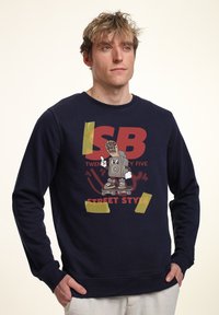 Felpa blu navy con una grafica audace di un personaggio dei cartoni animati con un boombox, accentuata da dettagli gialli in stile nastro. Materiale misto cotone.