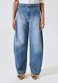 Jean large bleu clair, avec une coupe décontractée, une texture délavée, cinq poches et une fermeture à bouton. Associé à des chaussures dorées à bout pointu.