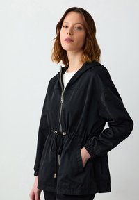 Zwarte lichtgewicht jas met een capuchon, verstelbare taille en gouden hardware. Voorzien van elastische manchetten en zijzakken. Gladde textuur.