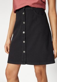 Zwarte denim rok met een knoopsluiting aan de voorkant, twee zakken en een gestructureerde vorm. De stof heeft een gladde textuur met subtiele stikdetails.