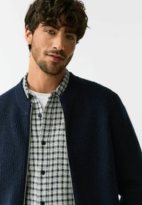 Cardigan bleumarin cu dungi, purtat peste o cămașă cu nasturi în carouri alb și albastru închis, cu decolteu rotund și textură subtilă.