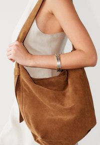 Sac bandoulière en daim marron avec un design souple et décontracté et une large sangle. Présente des coutures discrètes et une texture lisse.