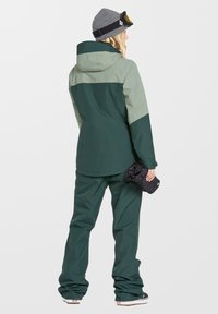 Grüne Skijacke mit hellgrünen Akzenten, ausgestattet mit Kapuze und verstellbaren Bündchen, kombiniert mit passenden grünen Skihosen. Hand in Hand mit gerollten Handschuhen.