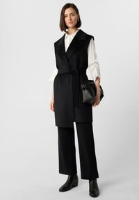 Schwarze ärmellose Weste mit einem Gürtel in der Taille, kombiniert mit einem weißen Hemd und weiten schwarzen Hosen. Glänzende schwarze Stiefeletten. Glatte, strukturierte Stoffqualität.