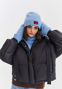 Čierna prešívaná bunda s krátkym strihom, nadrozmernými rukávmi a horizontálnym prešívaním. Mäkká svetlomodrá pletená čiapka a rukavice s textúrovaným povrchom.