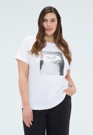 Kobieta o długich brązowych włosach, ubrana w białą koszulkę z czarno-białym zdjęciem kota, oczy kota zostały zastąpione narysowanymi wściekłymi oczami oraz czarne spodnie.