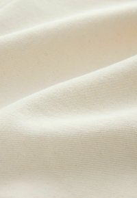 Gros plan sur un tissu doux de couleur crème avec une texture subtile en côtes diagonales et des plis délicats créant des jeux de lumière et d'ombre.