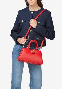Bolso de mano de cuero rojo con una forma estructurada, asas superiores dobles y una correa para el hombro desmontable. Los acentos de hardware dorado realzan el diseño.