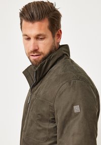 Grüne gesteppte Jacke mit hohem Kragen, Reißverschluss und strukturierter Stoffoberfläche. Mit einem Aufnäher-Logo auf der linken Schulter.