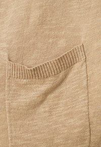 Tessuto lavorato a maglia beige con una tasca con dettaglio a coste. La texture è liscia con leggere variazioni di tonalità.
