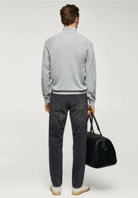 Uomo in piedi di spalle, che indossa un maglione grigio chiaro, jeans scuri, scarpe bianche, e tiene in mano destra una borsa da viaggio in pelle nera.