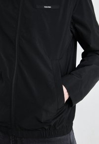 Črna zip-up jakna iz gladkega materiala, s stoječim ovratnikom, elastičnimi manšetami in stranskimi žepi. Logo na zgornjem levem predelu prsi.
