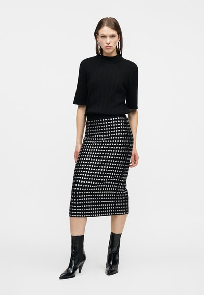 Γυναίκα που φοράει μαύρο τοπ με κοντά μανίκια, μαύρη μίντι φούστα με ασημένιο σχέδιο με οπές, μαύρα μποτάκια με τακούνι και μακριά σκουλαρίκια.