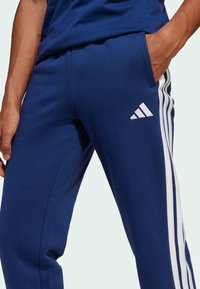 Pantalons de sport bleu marine en tissu doux, avec des accents blancs à trois rayures sur les côtés et un logo brodé sur la cuisse.
