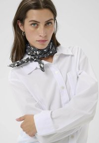 Camisa de algodón blanca con un frente con botones, acompañada de una bufanda negra con un patrón floral atada al cuello, mostrando una textura suave.