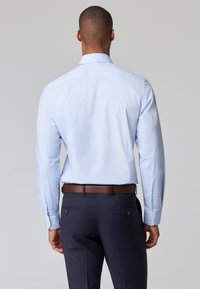 Camicia azzurra a maniche lunghe con colletto classico; materiale in misto cotone. Abbinata a pantaloni blu scuro e una cintura marrone. Dettagli a bottoni sui polsini.