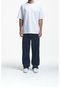 Camiseta blanca de gran tamaño hecha de algodón, combinada con jeans de mezclilla azul oscuro de corte holgado. El calzado son zapatillas deportivas gris claro con un diseño simple.