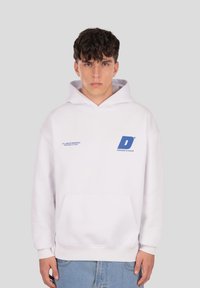 Biela mikina s kapucňou so zadným vreckom, s modrým textovým logom a značkovaním. Jemná látka, voľný strih s rebrovanými manžetami.