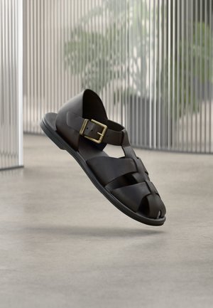 Sandalia masculina de cuero negro con tiras trenzadas y hebilla dorada, flotando sobre un suelo de hormigón liso en un ambiente interior moderno y minimalista.