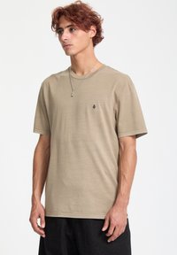 Beige bomulds T-shirt med rund hals og korte ærmer, prydet med et lille sort logo på venstre bryst. Glat tekstur, afslappet pasform.
