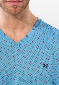 Hellblaues V-Ausschnitt-T-Shirt mit einem gemusterten Design aus roten Quadraten und schwarzen Punkten. Es hat ein kleines Logo auf der linken Seite.