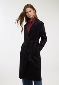 Cappotto nero con cintura, maniche lunghe, colletto con revers e tasche laterali, abbinato a una camicetta a righe rosse e bianche sotto.