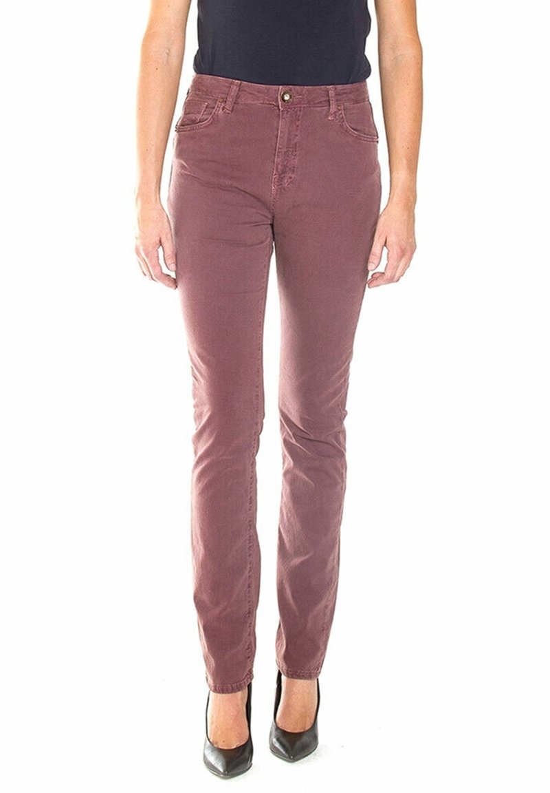 Femme portant un pantalon slim couleur rose poudré et des talons noirs à bout pointu, debout devant un fond blanc.
