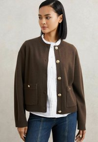 Rudas vilnonis kardiganas su apvalia iškirpte, auksiniais mygtukais, priekinių kišenių ir šonkaulinėmis rankovėmis, dėvimas virš baltos bluzkytės.