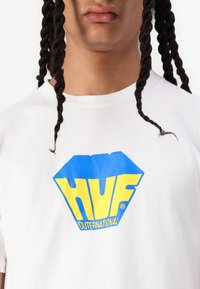 Hvid bomulds t-shirt med et stort blåt og gult grafisk logo "HUF OUTERNATIONAL" i en diamantform på brystet.