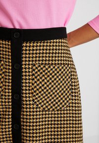 Jupe pied-de-poule noire et jaune avec une ceinture noire, dotée d'une fermeture à boutons et d'une poche latérale. Tissu texturé avec une forme structurée.