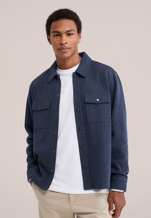 Jeune homme portant une veste bleu marine boutonnée sur une chemise blanche et un pantalon beige, debout devant un fond clair uni.