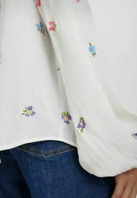 Blusa floreale bianca con maniche a sbuffo, caratterizzata da fiori ricamati di colore rosa, blu e viola su un tessuto testurizzato. Jeans in denim blu visibili.