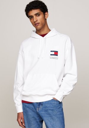 Sudadera blanca con bolsillo frontal, capucha ajustable y un logo con detalles en azul marino y rojo. Combinada con jeans de mezclilla azul.