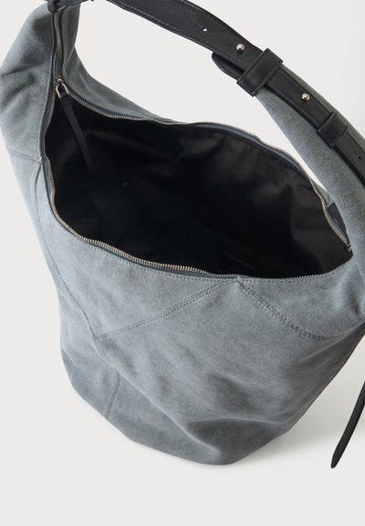 Sac d'épaule en tissu gris doux avec une bandoulière en cuir noir réglable et une fermeture éclair ouverte laissant voir la doublure intérieure noire.
