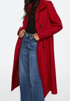 Femme portant un long manteau rouge par-dessus un haut noir et un jean bleu à taille haute et jambes larges, les mains dans les poches du jean.