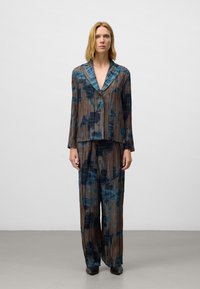 Vrouw in een losse, geplooide blouse met een bruin en blauw patroon en bijpassende wijde broek met lange mouwen, staand tegen een effen lichte achtergrond.