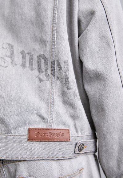 Lys grå denimjakke med et falmet "Palm Angels" logo, lædermærke og metalknapdetalje. Har stitchede sømme og en afslappet pasform.