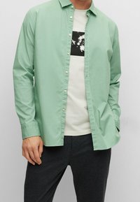 Camicia verde menta con bottoni in tessuto leggero, a maniche lunghe, indossata sopra una t-shirt grigia chiara con una stampa grafica nera.