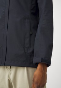 Veste imperméable navy à coupe droite, avec une poche frontale et une sangle Velcro au poignet ; texture lisse et design moderne.
