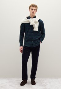 Geruit overhemd in donkergroen en marineblauw, gecombineerd met marineblauwe corduroy broek. Witte gebreide sjaal nonchalant over de schouders gedrapeerd. Bruine loafers.