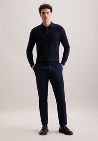 Mężczyzna stojący z rękami w kieszeniach, ubrany w granatową koszulę polo z długim rękawem, granatowe spodnie i czarne buty wizytowe, na jednolitym tle.